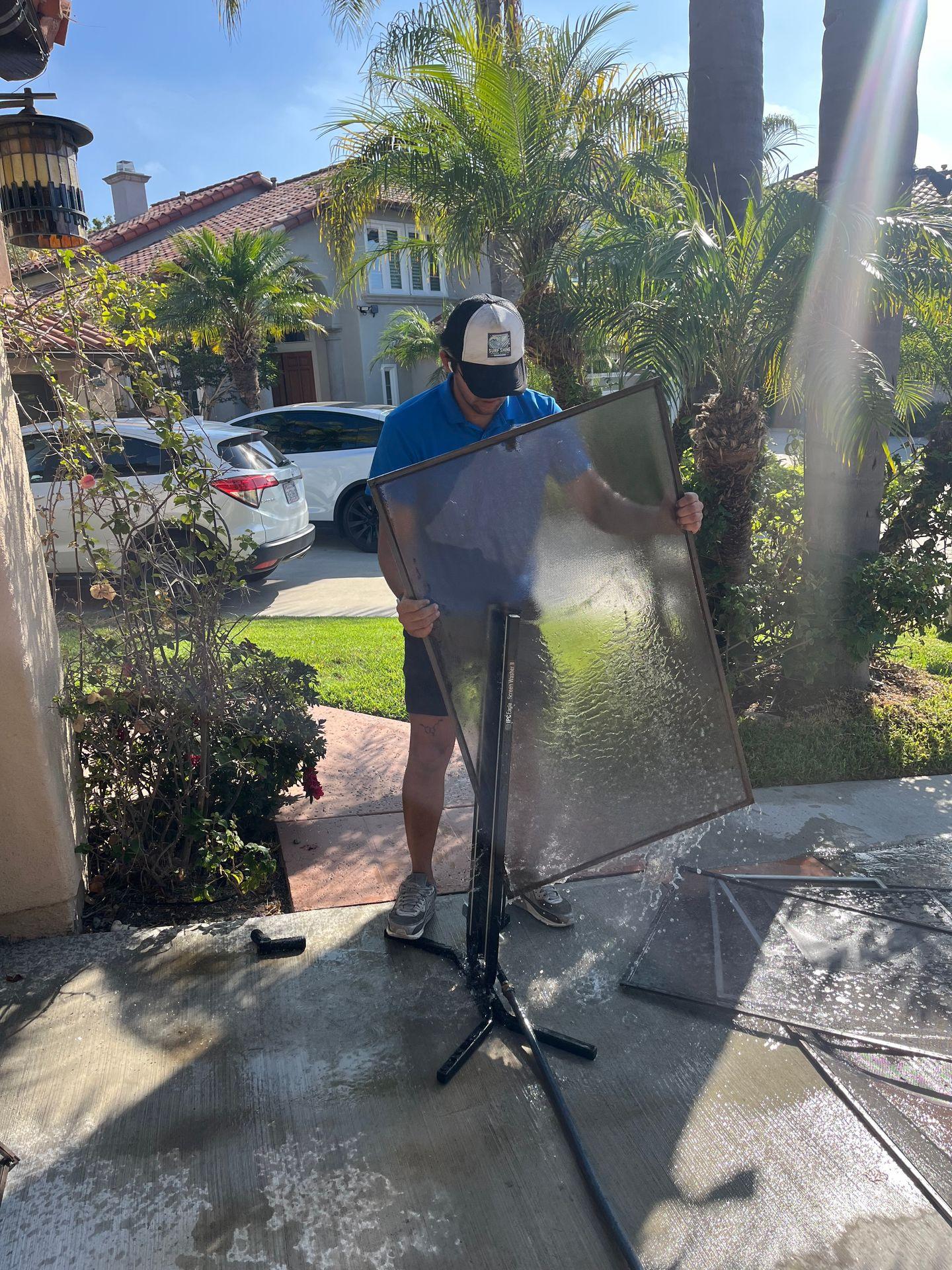 Person cleaning a glass panel outdoors near a garden with cars and houses in the background.