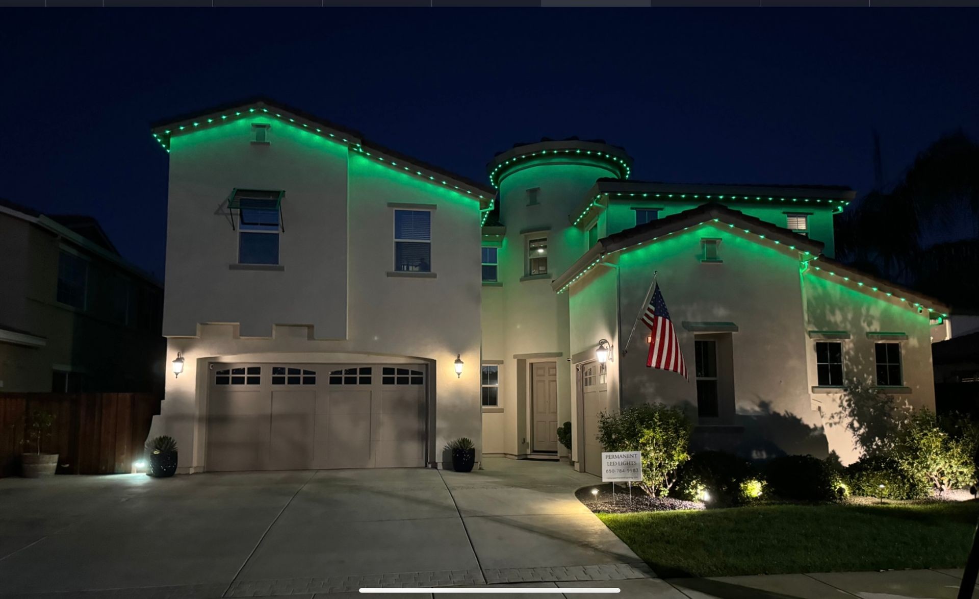 Two-story house with green LED lights, American flag, and a garage at night.